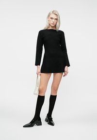 Nuori nainen, jolla on vaaleat hiukset, pukeutuneena mustaan pitkähihaiseen mini-mekkoon, polven yli ulottuviin mustiin sukkiin, mustiin korkokenkiin ja hänellä on pieni valkoinen käsilaukku.