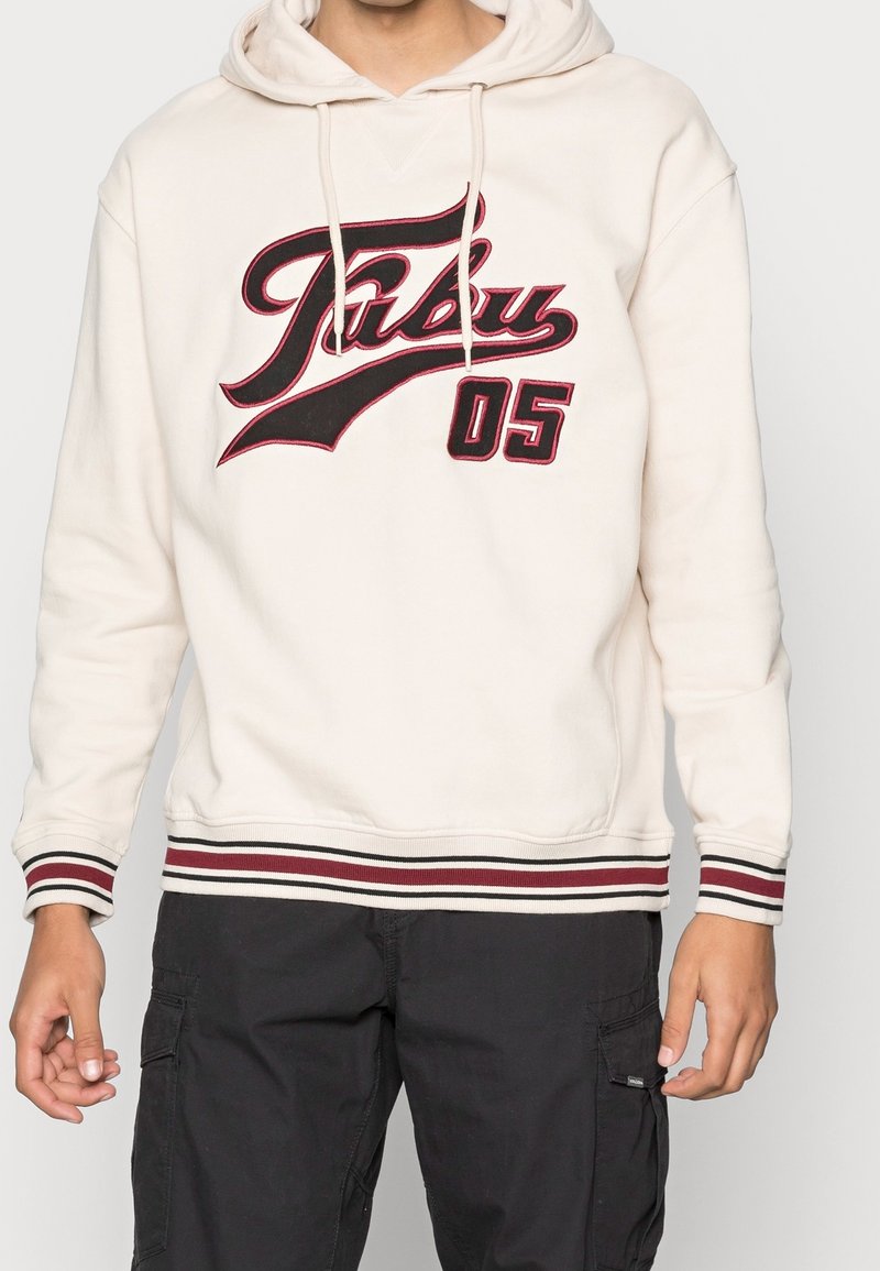 Roomkleurige hoodie met het rode en zwarte grafische logo "Fubu 05." Voorzien van een trekkoord in de capuchon en gestreepte manchetten met rode en zwarte banden. Zachte textuur.