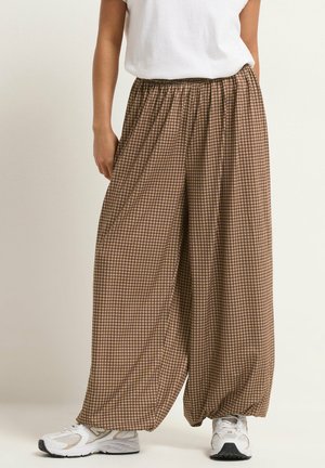 Person wearing loose brown and beige checkered pants with elastic ankles, white sneakers, and a white short-sleeve top.