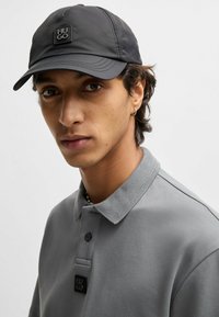 Sort kasket med et logo-patch, der har en glat og strømlinet finish. Sammen med en lys grå polo med krave og knapdetaljer.
