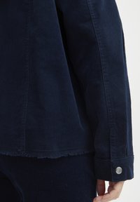 Giacca di velluto a coste blu navy con orlo sfrangiato, dotata di chiusura a bottone sui polsini e linee verticali testurizzate su tutto il tessuto.