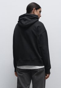 Felpa con cappuccio nera dal taglio rilassato, in tessuto morbido, con polsini a costine. Presenta una tasca a marsupio ed è indossata sopra una camicia bianca.