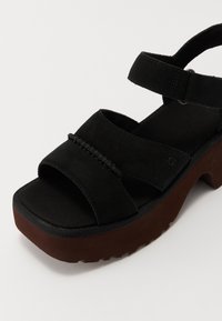 Zwarte suède plateau sandaal met twee brede banden, enkelband met gespsluiting en een dikke bruine zool met profiel.