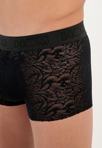 Schwarze Spitzen-Boxershorts mit elastischem Bund, versehen mit dem geprägten "HOM"-Logo. Der Stoff hat ein florales Muster und eine glatte Textur.