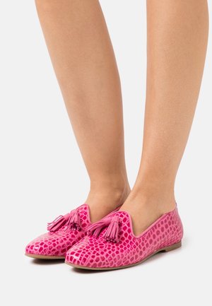 Füße, die leuchtend pinkfarbene, strukturierte Mokassins mit Quasten tragen, vor einem schlichten weißen Hintergrund.