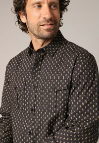 Zwarte overhemden met knopen met een schedelpatroon in goud, twee borstzakken en een klassieke kraag. De stoftekstuur lijkt soepel en lichtgewicht.