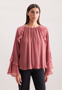 Blusa fluida di un rosa tenue con maniche a volant, texture plissettata e scollatura rotonda. Il tessuto sembra leggero e morbido.