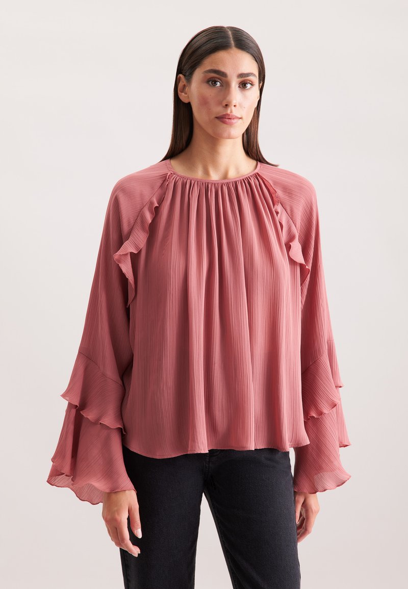 Blusa fluida di un rosa tenue con maniche a volant, texture plissettata e scollatura rotonda. Il tessuto sembra leggero e morbido.