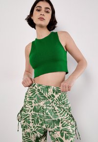 Le modèle porte un débardeur vert court et un pantalon beige à motifs de feuilles vertes, faisant face à la caméra avec une expression confiante.