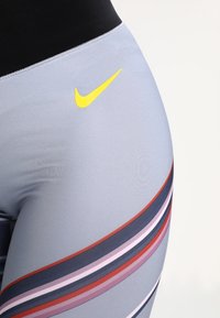 Legginsy Nike w jasnoszarym kolorze z czarnym pasem, z wielokolorowymi poziomymi paskami i detalem w postaci żółtego logo Swoosh.