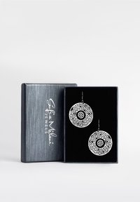 Runda silverörhängen med intrikata utskurna mönster och en solid svart mitt, visade i en mörk sammetsfodrad presentlåda.