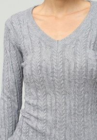 Pull en tricot gris avec un col en V, des motifs de torsades complexes et des manches longues. La texture semble douce et confortable.
