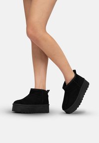 Botas de tobillo de ante negro con suela de plataforma gruesa, punta redondeada y adornos cosidos. Incluyen una tirador en la parte trasera para ponerlas con facilidad.