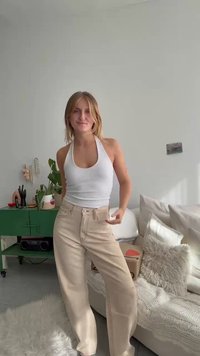 Top halter branco e calças largas bege, com uma cintura alta e costura visível. A cena inclui um armário verde e móveis suaves.