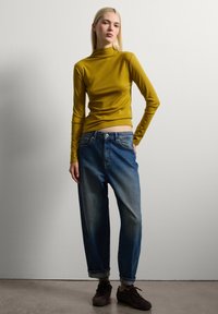 Gele langemouwen top met een hoge hals, gecombineerd met losse, lichtblauwe spijkerbroeken. Model staat op een neutrale achtergrond en draagt donkere schoenen.