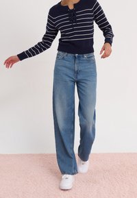 Marinrandig tröja med en knytdetalj, ihop med ljusblå vida jeans och vita sneakers, mot en mjuk rosa bakgrund.