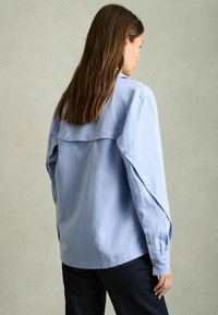 Camisa de manga larga azul claro de algodón, con botones a presión en las mangas y un diseño en la parte posterior con capas. Llevada con jeans oscuros.