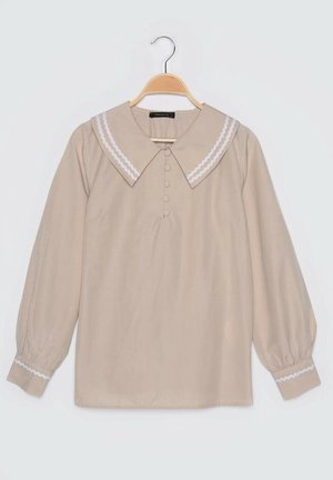 Blusa beige de manga larga con cuello grande y puntiagudo con ribete blanco y botones cubiertos de tela en la parte delantera, exhibida en una percha de madera.