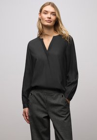 Maglia nera con scollo a V e maniche lunghe; realizzata in tessuto liscio con una texture sottile. Abbinata a pantaloni grigi a fantasia con bottoni.