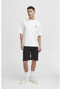 Hvid bomulds T-shirt med korte ærmer og blåt blomstermønster på venstre bryst, parret med sorte shorts og hvide sneakers.