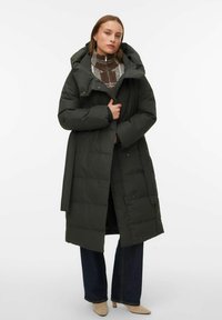 Tumši zaļa pufi jaka ar kapuci, ar šķindīgu dizainu, aizvēramu rāvējslēdzēju un sānu kabatām, uzvilkta pār rakstainu džemperi un zilām džinsām.