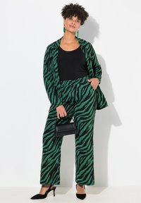 Grön och svart zebra-mönstrad kostym med långa ärmar och vida byxor. Modellen håller en svart väska och bär svarta skor och gröna örhängen.