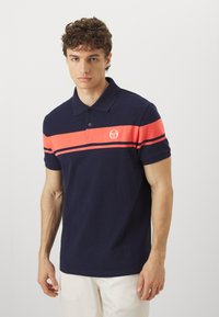 Námornícky modré polo tričko vyrobené z bavlny, s koralovou horizontálnou páskou a logom. Krátke rukávy a klasický dizajn goliera.