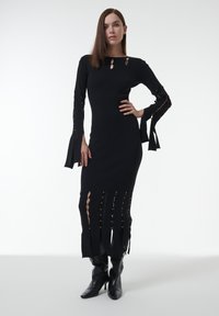 Schwarzes Strickkleid mit langen Ärmeln, das Ausschnitte und goldene Akzente aufweist, körperbetonte Silhouette und einen Saum mit Fransenverzierung hat. Kombiniert mit schwarzen Stiefeln.