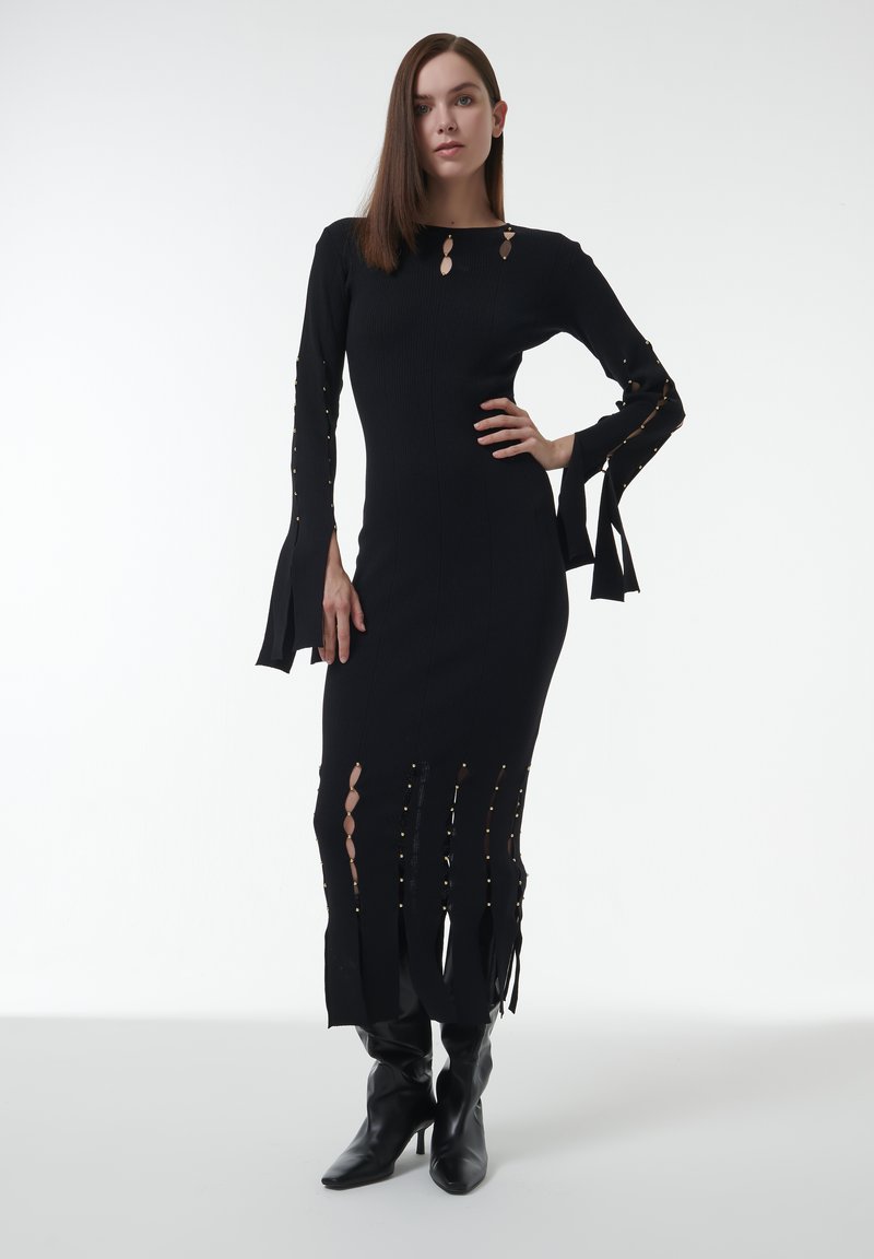 Schwarzes Strickkleid mit langen Ärmeln, das Ausschnitte und goldene Akzente aufweist, körperbetonte Silhouette und einen Saum mit Fransenverzierung hat. Kombiniert mit schwarzen Stiefeln.