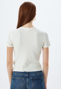 Ribbet, kortærmet t-shirt i off-white, med rund halsudskæring og en tætsiddende silhuet. Bæres med højtaljede blå jeans.