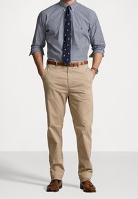 Mężczyzna w niebiesko-białą w kratkę koszuli z podwiniętymi rękawami, granatowym krawacie z emblematem, beżowych spodniach, brązowym skórzanym pasku, zegarku i brązowych mokassynach z frędzlami.