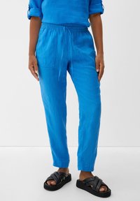 Blå linnebyxor med elastisk midja och dragsnöre, med två sidofickor, tillsammans med svarta slidesandaler.