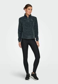 Tamni flis pulover s ovratnikom na zip, sa strukturalnim leopard uzorkom, u kombinaciji s crnim tajicama koje imaju suptilan uzorak, nošene s tenisicama.