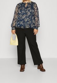 Blusa floreale blu navy con maniche lunghe trasparenti, dettaglio di scollatura a chiave, abbinata a pantaloni neri a gamba larga e stivali con tacco marroni, mentre tiene una borsa gialla.