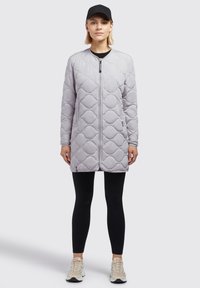 Manteau long matelassé gris clair avec une fermeture éclair à l'avant, un col rond et des poches latérales. Associé à des leggings noirs et des baskets beiges.