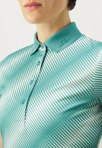 Teal krāsas polo krekls ar baltām diagonālām svītrām, aprīkots ar pogāmu priekšpusi, strukturētu apkakli un teksturētu logo detaļu uz apkakles.