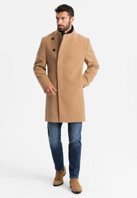 Beiger Wollmantel mit hohem Kragen und asymmetrischem Verschluss, ausgestattet mit zwei schwarzen Knöpfen. Getragen über blauen Jeans und tanfarbigen Wildlederstiefeln.