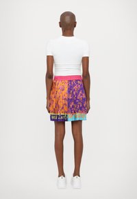 Gonna plissettata colorata con sezioni arancioni e viola, caratterizzata da un design a motivo e una fascia in vita rosa con testo. Top e scarpe bianchi visibili.