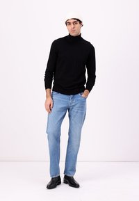 Suéter negro con cuello alto, jeans azul claro y botas negras hasta el tobillo. El modelo está de pie contra un fondo blanco, usando un gorro blanco.
