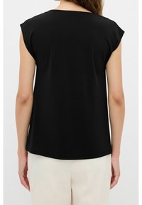 Mujer vestida con una blusa sin mangas de color negro combinada con pantalones beige claros, vista desde atrás contra un fondo simple.