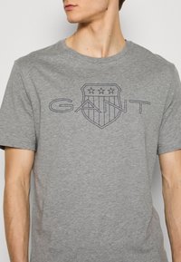 T-shirt gris en coton avec un logo bouclier sombre comportant trois étoiles et le mot "GANT" en lettres majuscules. Manches courtes et col rond.