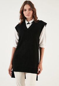 Black sleeveless sweater vest with a v-neckline, ribbed hem, and slits on the sides, worn over a white collared shirt. Soft knit material.