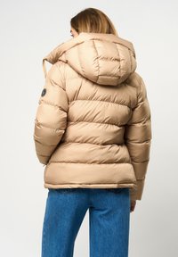 Béžová puffer bunda s kapucňou, so zafúkaným dizajnom, hrubým polstrovaním a logom na rukáve. Nosené so modrými džínsami.