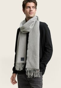 Grijze geweven sjaal met franjes, gemaakt van zacht materiaal. Weergegeven op een model dat een zwart shirt draagt tegen een neutrale achtergrond.