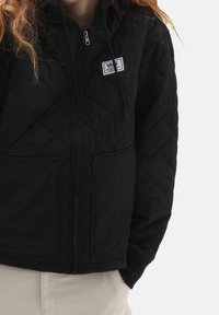 Person som bär en svart quiltad zip-up hoodie med framfickor och en Vans-logotyp på bröstet, i kombination med ljusbeiga byxor.