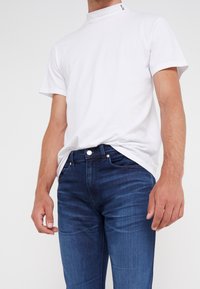 Camiseta de algodón blanca con cuello alto, combinada con jeans de mezclilla ajustados de color azul oscuro que presentan un diseño clásico de cinco bolsillos y detalle de botón de metal.