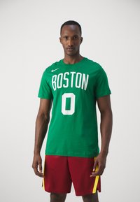 Grøn bomulds T-shirt med "BOSTON" og "0" trykt i hvid, med rund hals og korte ærmer. Bæres med røde shorts med gule detaljer.