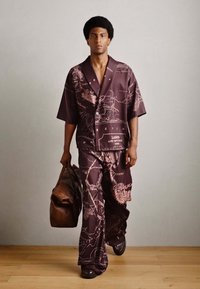 Lila Hemd mit Knöpfen und passende weit geschnittene Hose mit einem Kartendesign, kombiniert mit einer braunen Ledertasche und schwarzen Schuhen.