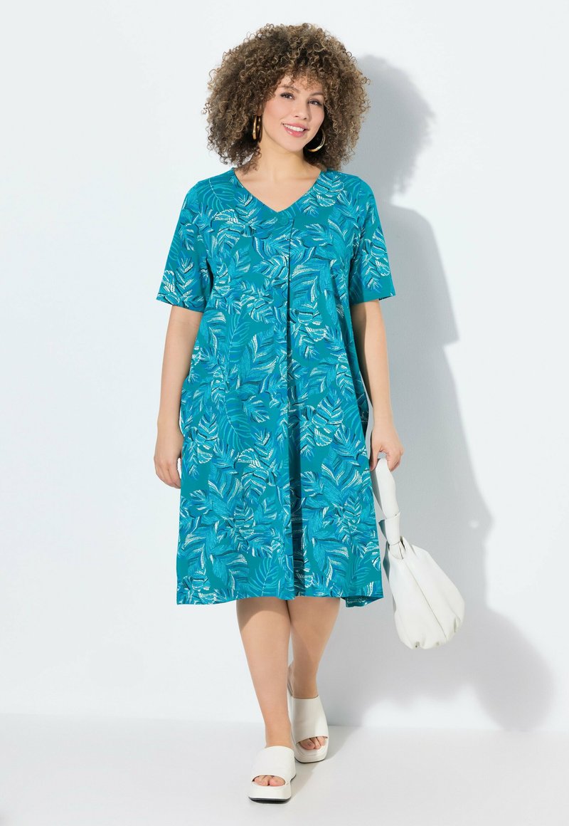 Vestido verde azulado con estampado floral; mangas cortas, escote en forma de V, corte holgado. Combinado con sandalias de plataforma blancas y un bolso blanco.