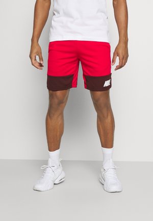 Atlet iført røde shorts med mørkebrune lommer og hvidt Nike-logo, hvide sokker, hvide sneakers og en hvid t-shirt mod en grå baggrund.